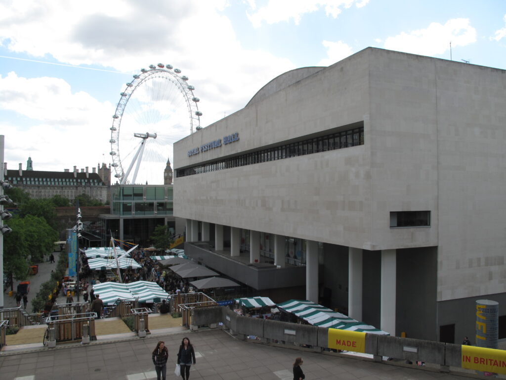 "Royal Festival Hall, Southbank Centre” by duncan c; gray rectangular building with columns on first level overlooks a fair with striped tents and a ferris wheel.