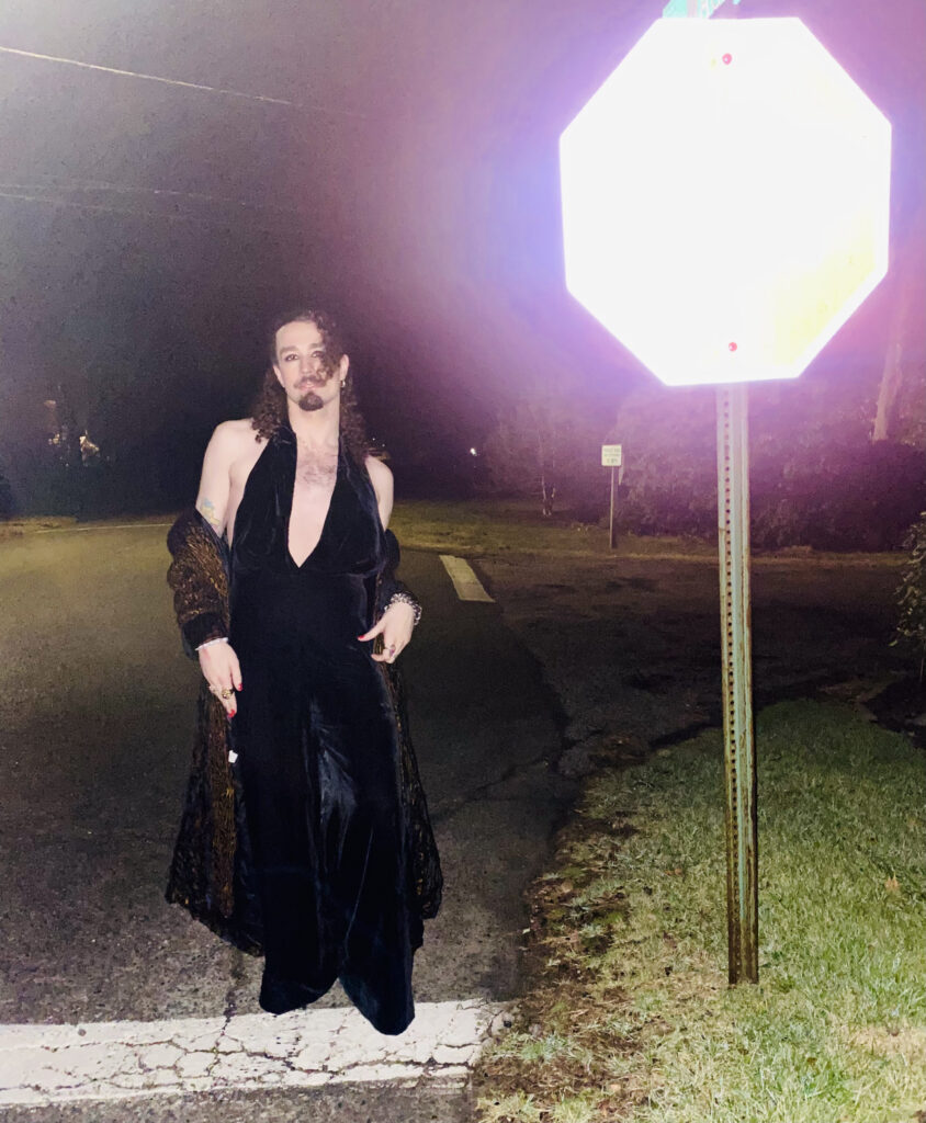 Caelan Ernest (cyborg?) in a velvet jumpsuit and long coat off the shoulders standing next to a stop sign washed out in reflection. It's night time on a residential street.
